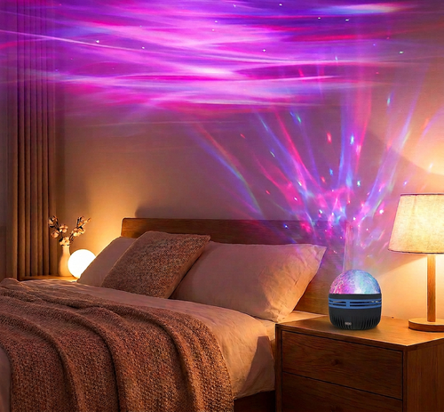 A small blue projector lamp sits on a wooden bedside table in a bedroom, casting colorful light beams across the wall above a bed with pillows and a blanket.